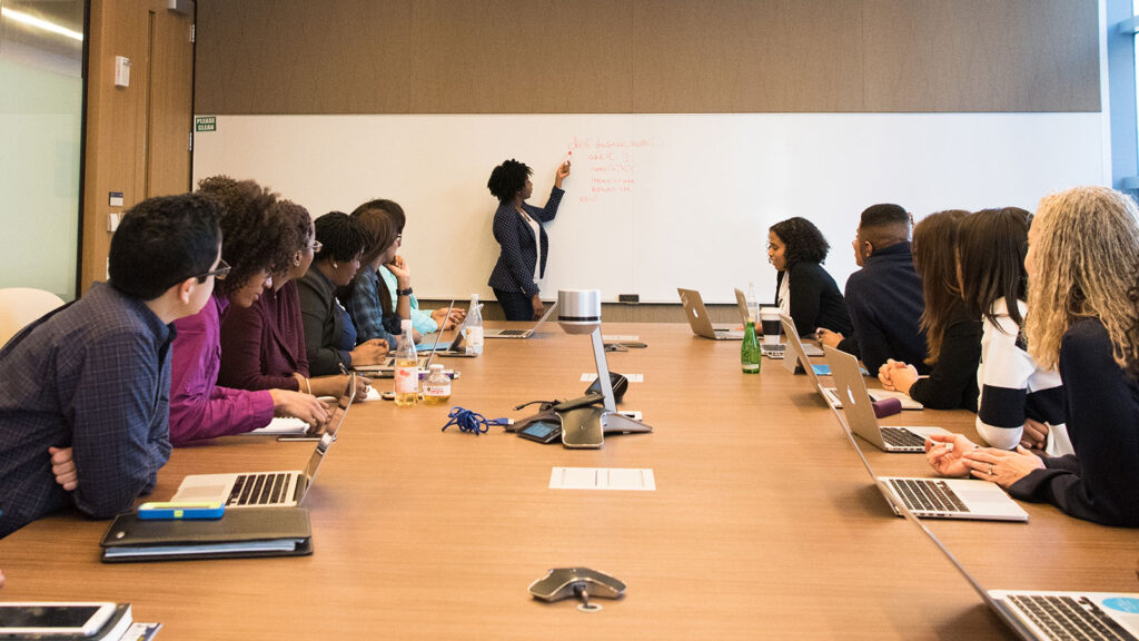 Women pointing to whiteboard with group of students at table with laptops listening. Representing a visual of a library career.